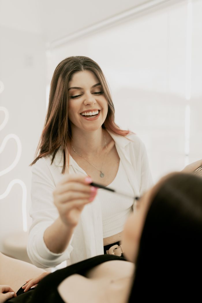 A makeup artist smiling while applying eyeshadow to a client in a well-lit beauty studio.