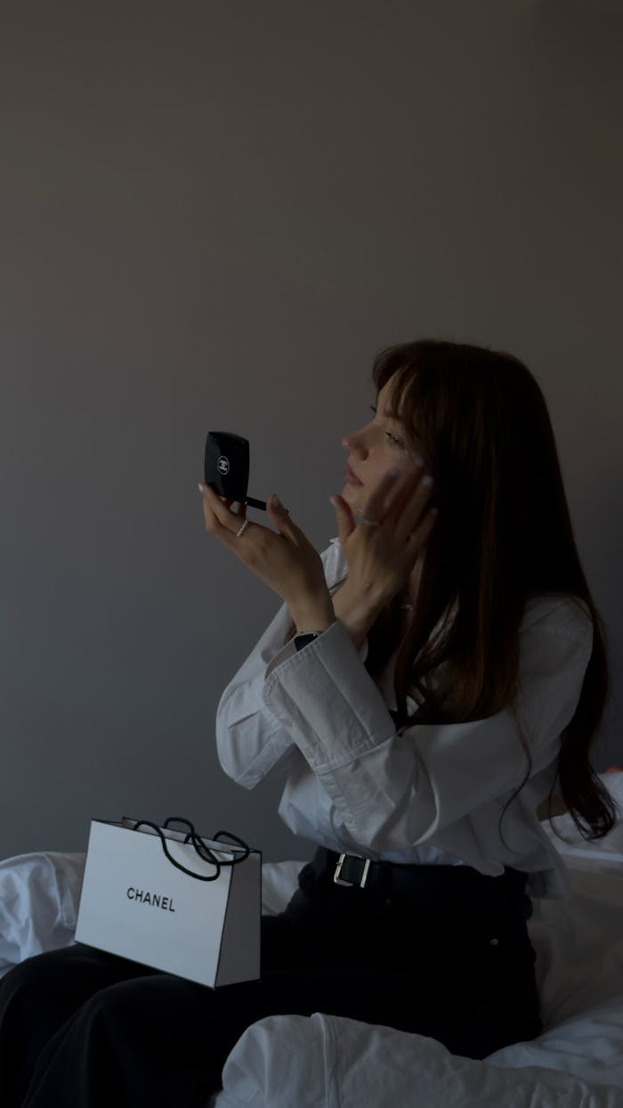 Adult woman applies makeup using a compact mirror, seated indoors with a shopping bag.